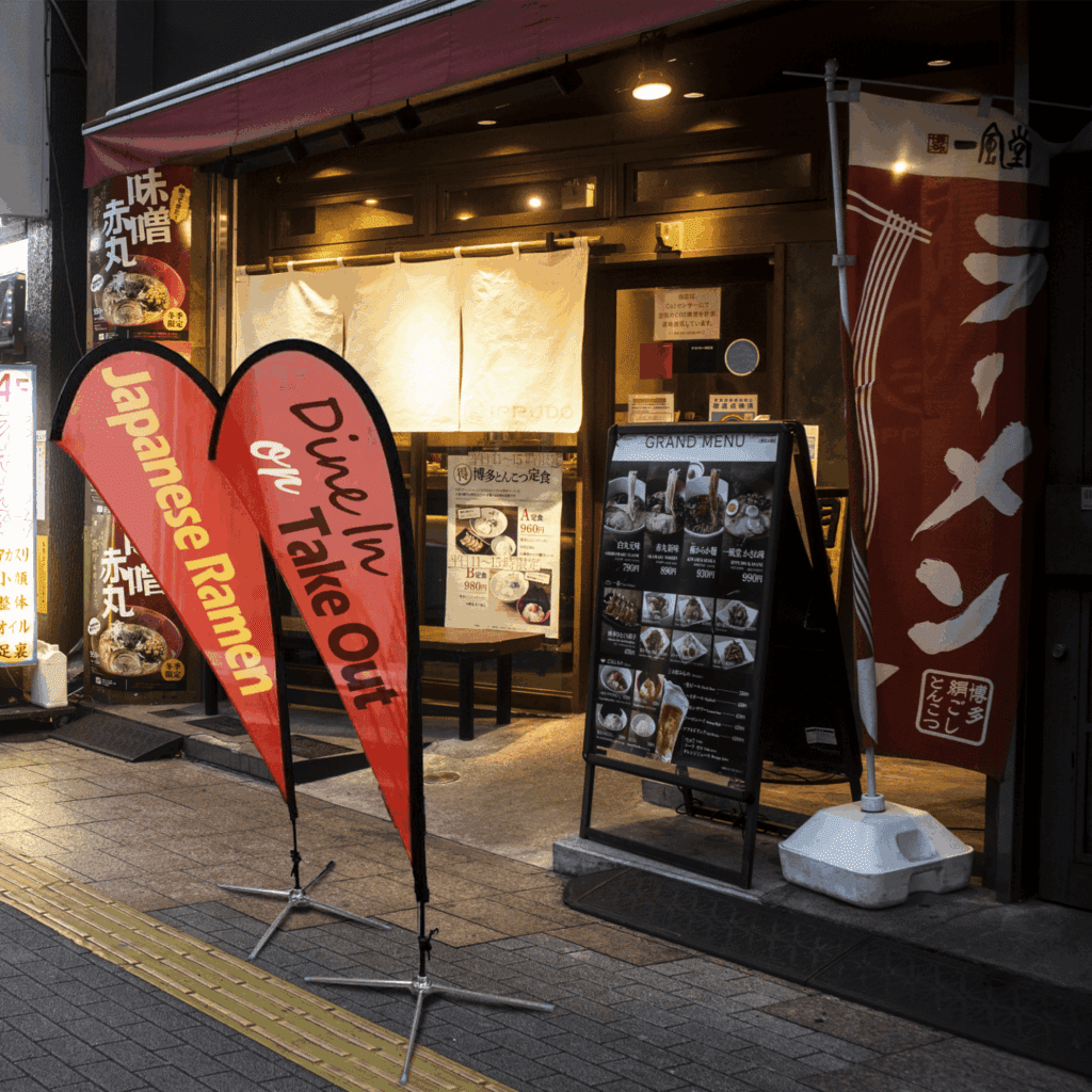 Custom teardrop flags outside a Japanese ramen restaurant at night.